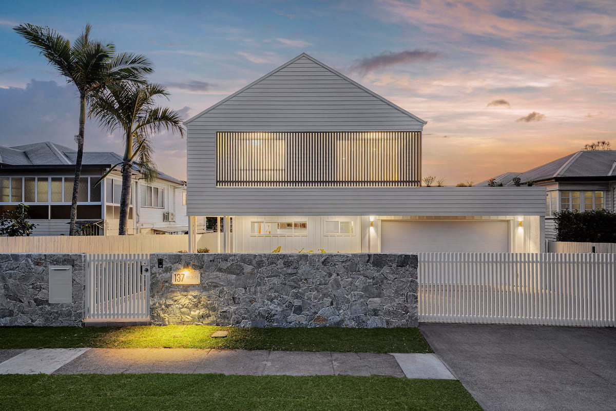White gabled Hamptons home at dusk with stone front wall and picket fence